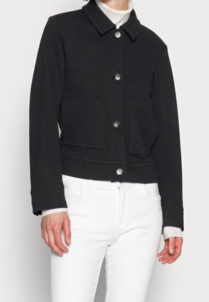 Femme portant une veste noire boutonnée sur un pull à col roulé blanc et un pantalon blanc, montrée du cou jusqu'aux hanches.