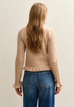Pull beige en tricot avec un design court, des manches texturées et un ourlet à volants, associé à un jean en denim bleu taille haute.