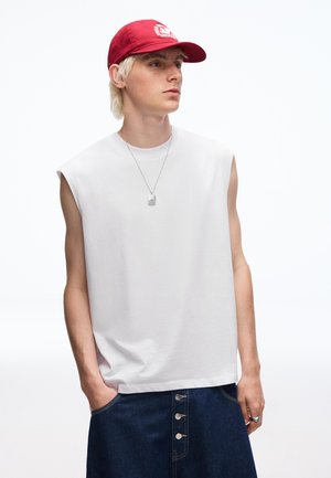 Fiatal személy ujjatlan fehér felsőben, piros baseballsapkában, ezüst nyaklánccal és sötétkék, gombos nadrágban, kezét zsebében tartva.