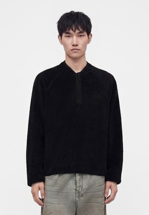 Jeune homme portant un pull polaire noir avec une fermeture éclair quart de tour, debout devant un fond blanc uni.