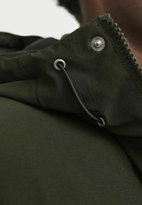 Oliivroheline kapuutsiga jakk sileda tekstuuriga, hõbedase tõmbluku ja kise detailidega, reguleeritava paelaga ning lähedalt vaade kraele.