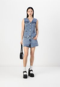 Denimvest med spiss kant, fem knapper, to frontlommer, sammen med denimshorts. Modellen holder en liten svart veske og har på seg platåsko.