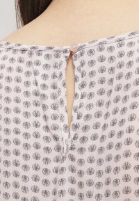 Tejido de color rosa claro con un diseño estampado de formas florales en gris. Presenta una abertura en forma de llave en la parte posterior, asegurada con un botón redondo.