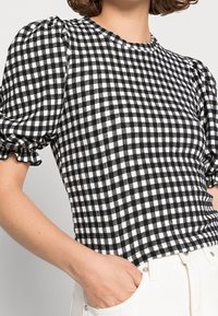 Top corto a maniche corte in check bianco e nero con bordi arricciati, scollatura rotonda e tessuto strutturato. Il modello ha una mano in tasca dei pantaloni bianchi.