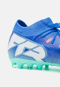 Zapatilla de fútbol azul con un upper sintético texturizado, marca blanca y una suela azul clara. Presenta cordones y un diseño de corte bajo.