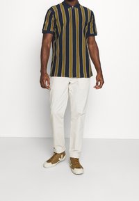 Kortärmad polo med vertikala ränder i marinblått, gult och vitt. Kombineras med krämfärgade byxor och bruna sneakers med vita accenter.