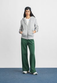 Jauna sieviete stāv ar rokām kabatās, ģērbjus pelēkā kapucē, zaļos treniņbiksēs, baltās kedās un melnā atpakaļgaitā cepurē uz zilas grīdas.