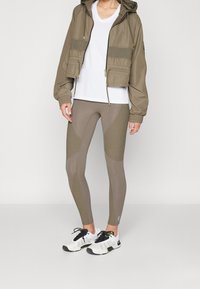 Chaqueta de oliva recortada con bolsillos frontales, camiseta blanca de cuello en V, y leggings ajustados color beige con paneles texturizados. Zapatillas deportivas negras con acentos blancos.