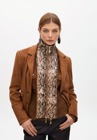 Žena s hladce učesanými vlasy má na sobě velké černé a zlaté náušnice, hnědou bundu přes top se vzorem hadí kůže na zip a tmavé kalhoty.