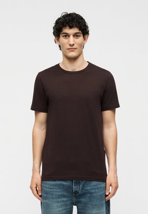 Νέος άνδρας με σγουρά σκουρόχρωμα μαλλιά, που φοράει ένα απλό κοντομάνικο μαύρο μπλουζάκι και μπλε τζιν, στέκεται μπροστά σε ένα απλό λευκό φόντο.