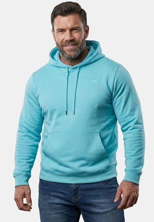 Man met baard, die een lichtblauwe hoodie en donkerblauwe spijkerbroek draagt, staand tegen een effen achtergrond, licht glimlachend.