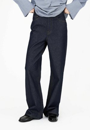 Persoon draagt donkere wijde jeans, zwarte schoenen en een blauw geruit overhemd met lange mouwen en uitlopende manchetten, staand met handen in de heupen.