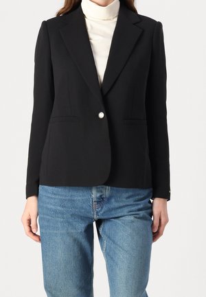 Vrouw draagt een nauwsluitend zwart colbert met een enkele zilveren knoop, een witte coltrui en blauwe spijkerbroek, staand tegen een effen achtergrond.