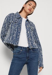 Denim jakna u plavoj boji s rastrganim bijelim detaljima, s ovratnikom, prednjim džepovima i gumbima. Uparena s tamnoplavim uskim trapericama.