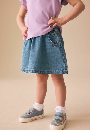 Dieťa v džínsovej sukni, fialovom tričku, bielych ponožkách s volánmi a modrých teniskách s výšivkou dúhy a kvetín, stojace s rukami v bok.