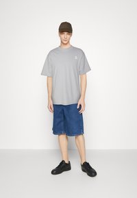 Grå t-skjorte med en liten hvit logo, kombinert med blå denimshorts. Modellen har på seg svarte joggesko og en olivengrønn caps.
