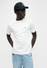 Λευκό βαμβακερό T-shirt με κοντά μανίκια, στρογγυλή λαιμόκοψη, με μικρό μαύρο λογότυπο στο στήθος. Συνδυασμένο με ανοιχτό μπλε τζιν και μαύρο καπέλο.