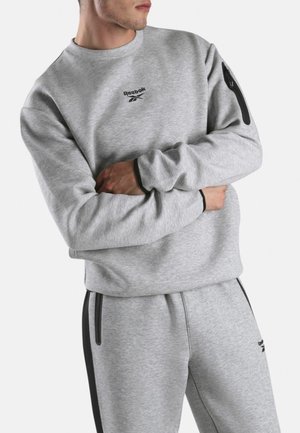 Homme portant un sweat-shirt Reebok gris clair et un pantalon de survêtement assorti avec des détails de fermeture éclair noirs, bras croisés sur fond blanc.