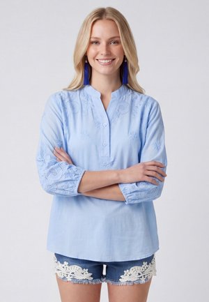 Vrouw glimlachend met gekruiste armen, draag een lichtblauw geborduurd bloesje, donkerblauwe oorbellen met kwastjes en denim shorts met witte kanten rand.