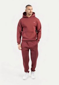 Sudadera con capucha color granate con bolsillo frontal y capucha ajustable, combinada con pantalones jogger ajustados a juego. Zapatillas blancas completan el conjunto. Textura suave.