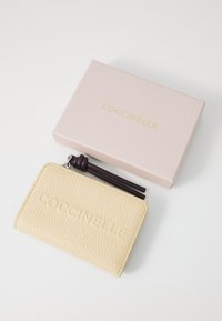 Portefeuille Coccinelle en cuir texturé beige, avec un logo embossé, accompagné d'une boîte rose avec des lettres dorées.