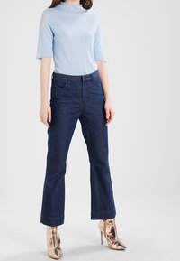 Ljusblå polotröja, mörk denimjeans med medelhög midja och utsvängda ben, samt metalliska roséguld högklackade skor. Slät textur, avslappnad design.