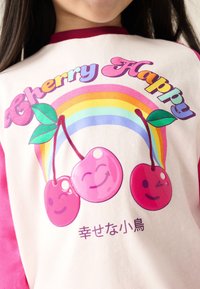 Valge särk roosade varrukatega, millel on värviline graafika kolmest naeratavast kirssist, vikerkaarest ja mängulise kirjaga tekst "Cherry Happy".