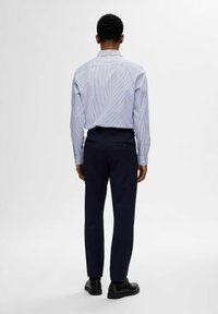 Vista posteriore di un uomo in camicia a righe azzurre chiare e pantaloni navy, con colletto standard, maniche lunghe e scarpe nere.