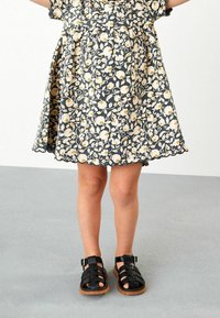 Ein Kind trägt ein dunkles Blumenkleid mit gewelltem Saum und schwarze Schnallsandalen. Es steht auf einem grauen Boden vor einem weißen Hintergrund.