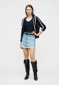 Γυναίκα με μαύρη ανοιχτή ζακέτα πλεκτή, μαύρη αμάνικη μπλούζα, ανοιχτόχρωμη φθαρμένη τζιν φούστα, μαύρη ζώνη και μαύρα μπότες μέχρι το γόνατο, στέκεται με το χέρι στη μέση.