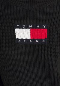 Svart stickad tyg med en rektangulär lappar som har Tommy Jeans-logotypen i vit, röd och marinblå garn.
