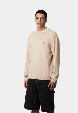 Suéter de punto beige con cuello redondo y puños acanalados, combinado con pantalones cortos de mezclilla negros que cuentan con grandes bolsillos y un ajuste relajado.