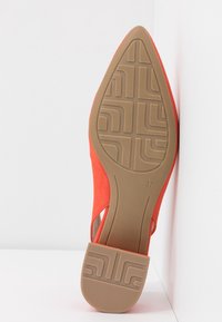 Chaussure plate orange à bout pointu montrant une semelle en caoutchouc marron à motifs et la taille 37, placée verticalement contre un coin blanc.