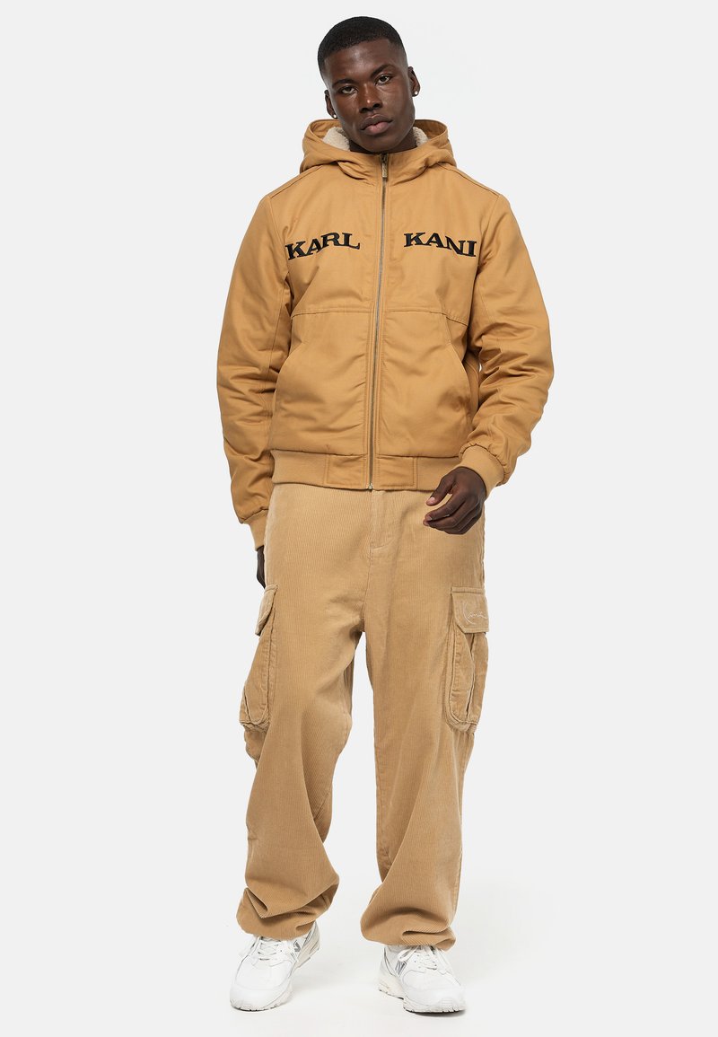 Tan bomber jacket with a hood, featuring black "KARL KANI" text on the front. Paired with beige corduroy cargo pants and white sneakers.