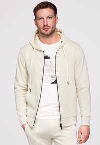 Homme portant un sweat à capuche zippé blanc cassé et un t-shirt blanc avec un motif graphique, les mains dans les poches, debout devant un fond clair uni.