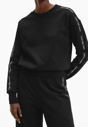 Sweat-shirt noir à manches longues et pantalon avec "Calvin Klein Jeans" imprimé à plusieurs reprises le long des manches et des côtés du pantalon, porté par une personne.