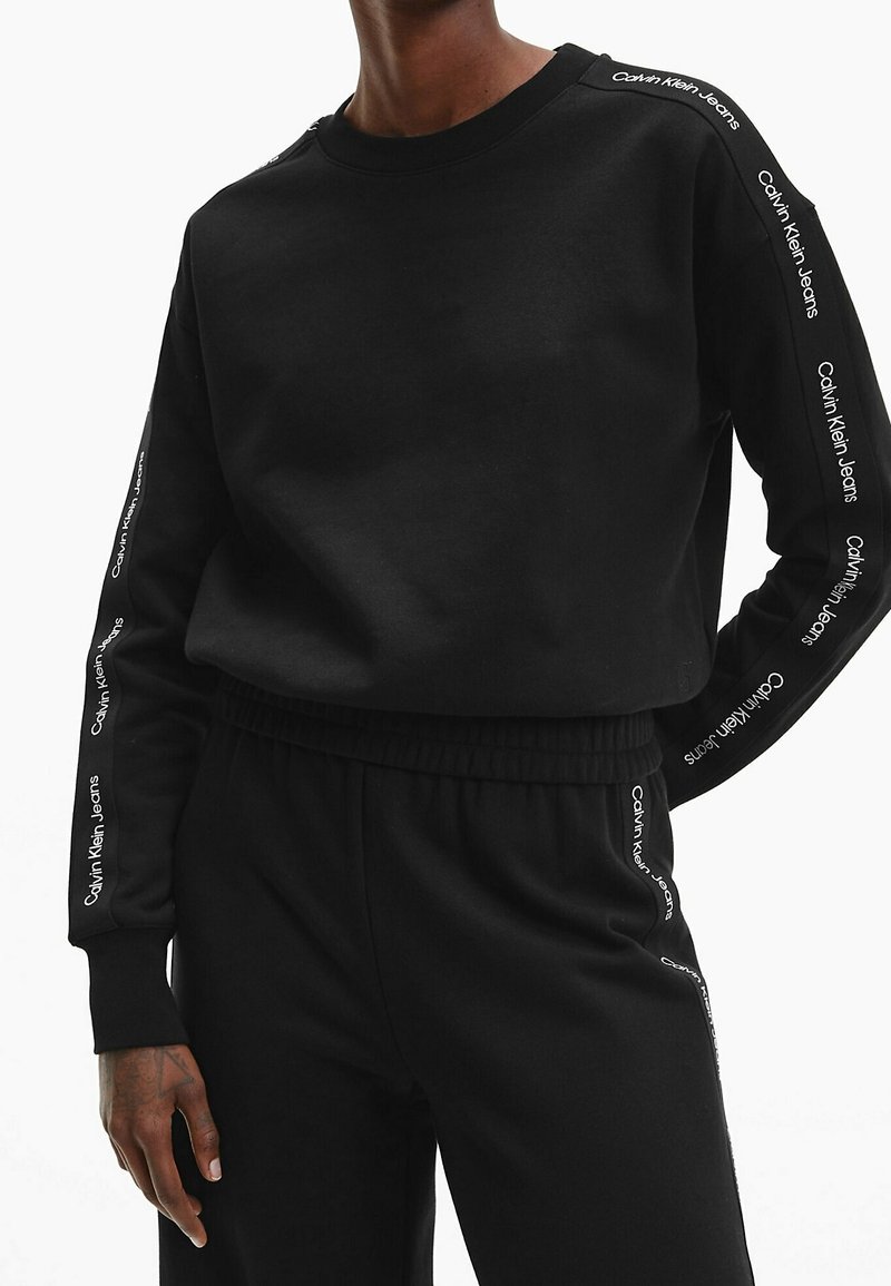 Sweat-shirt noir à manches longues et pantalon avec "Calvin Klein Jeans" imprimé à plusieurs reprises le long des manches et des côtés du pantalon, porté par une personne.