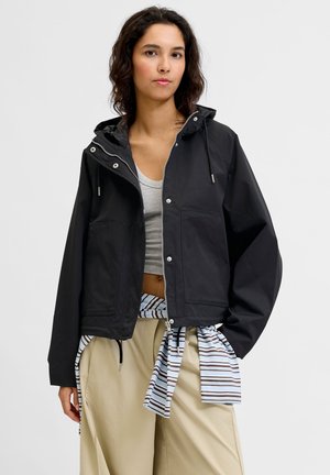 Mladá žena nosí čiernu kapucňovú bundu cez sivý crop top, béžové nohavice a pruhovanú košeľu zviazanú okolo pása.