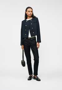 Marineblå kort jakke med gullknapper, kombinert med mørke jeans og svarte loafers. Modellen holder en liten svart håndveske. Enfarget bakgrunn.