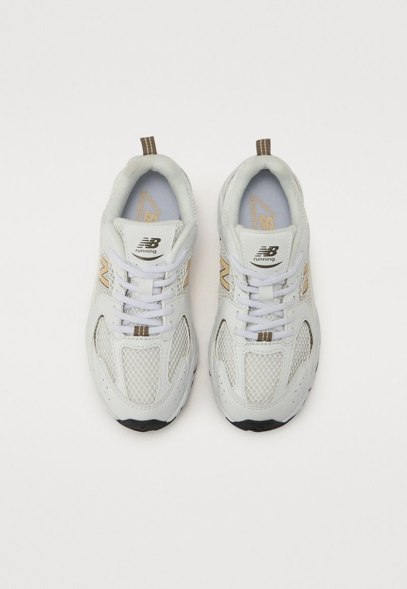 Witte mesh hardloopschoenen met gouden accenten, zwarte zolen en bruine treklussen. Beschikt over een gewatteerde enkelsteun en een vetersluiting.