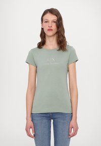 Lichtgroene t-shirt met korte mouwen, gemaakt van zachte katoen, met een logo met zilveren accenten over de borst. Gekleed met strakke blauwe jeans.
