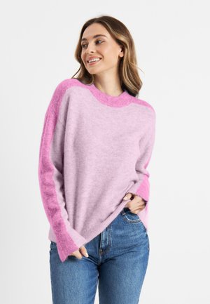 Woman smiling and looking left wearing a fuzzy pink and light purple sweater and blue jeans against a plain white background.