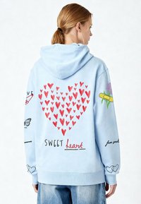 Sweat à capuche bleu clair en tissu doux avec un grand motif de cœur rouge, des éléments dessinés à la main, et des accents de texte au dos et sur les manches.