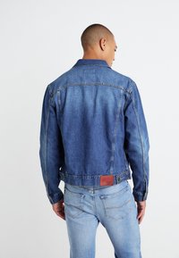 Jeansjakke i medium blå med knappelukking på kragen, lommer på siden og lærpatch-detalj på baksiden. Klassisk tettsittende design.