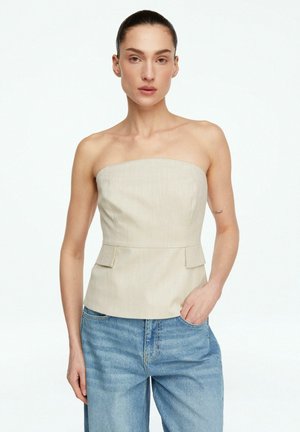 Kobieta o ciemnych włosach w beżowym topie bez ramiączek i niebieskich dżinsach pozująca z jedną ręką w kieszeni na białym tle.