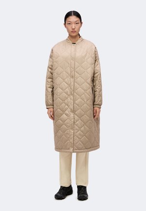 Person står vendt fremad iført en lang beige quiltet jakke, lyse bukser og sorte sko på en ensfarvet lys baggrund.