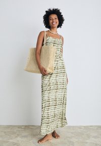 Maxi dress with green and beige horizontal tie-dye pattern, thin straps, and a fitted waist. Woven tan tote bag with fringed edges.
