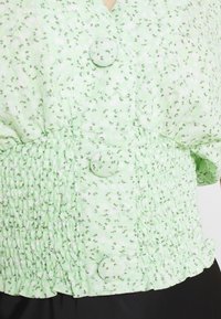 Top floreale verde con scollo a V e tre bottoni, caratterizzato da un design arricciato in vita e da un tessuto morbido e leggero.