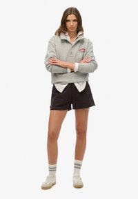 Szary sweatshirt z czerwonym logo "Superdry", nałożony na biały shirt z kołnierzykiem, w połączeniu z czarnymi szortami i paskowanymi skarpetkami. Białe sneakersy dopełniają całość.