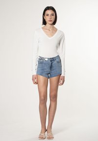 Weißes Langarmshirt mit V-Ausschnitt, kombiniert mit blauen Jeansshorts. Die Shorts haben einen ausgefransten Saum und auffällige Beschläge am Hosenbund.